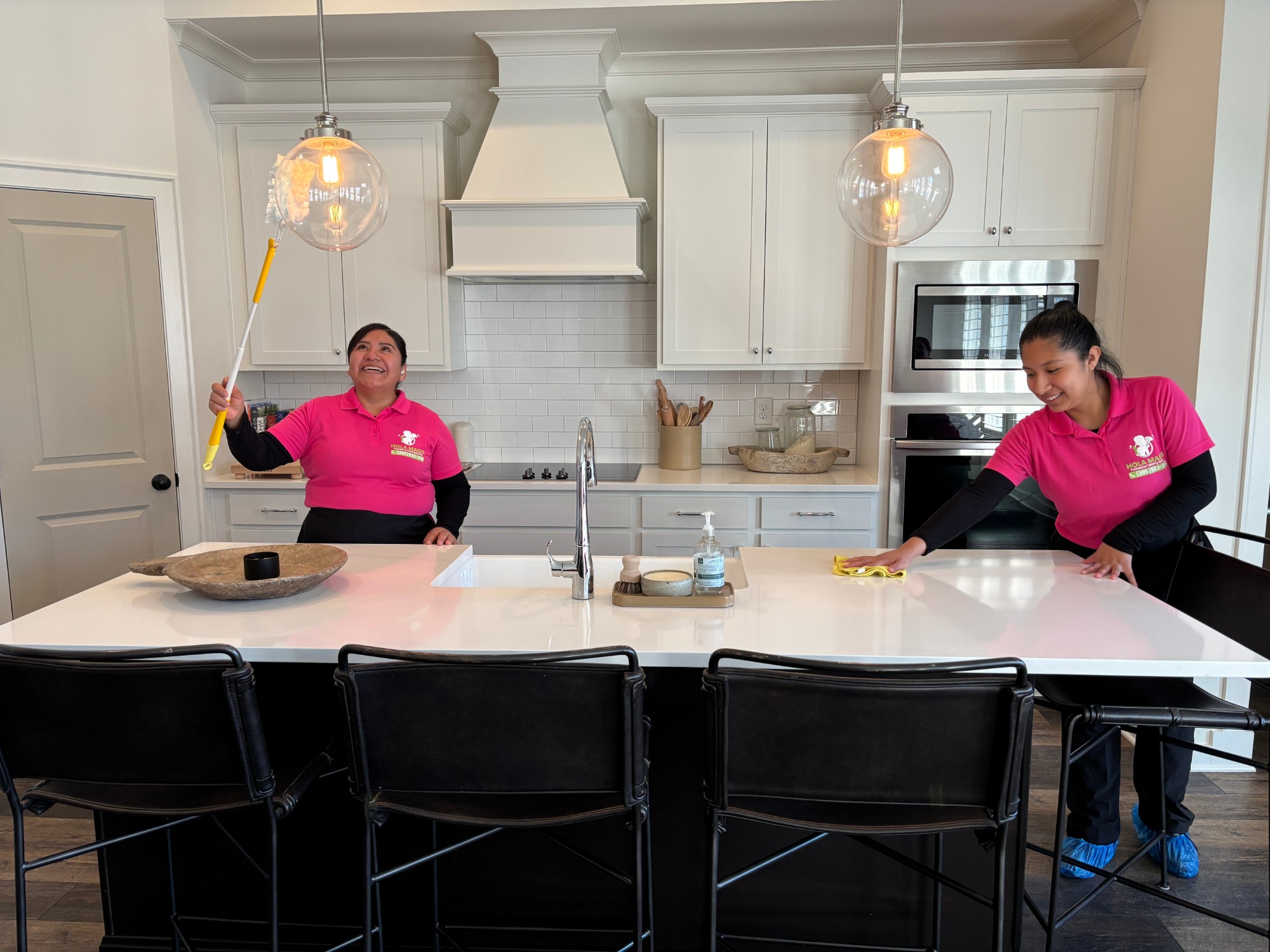 Two Hola Maids cleaners in pink uniforms cleaning a bright white kitchen